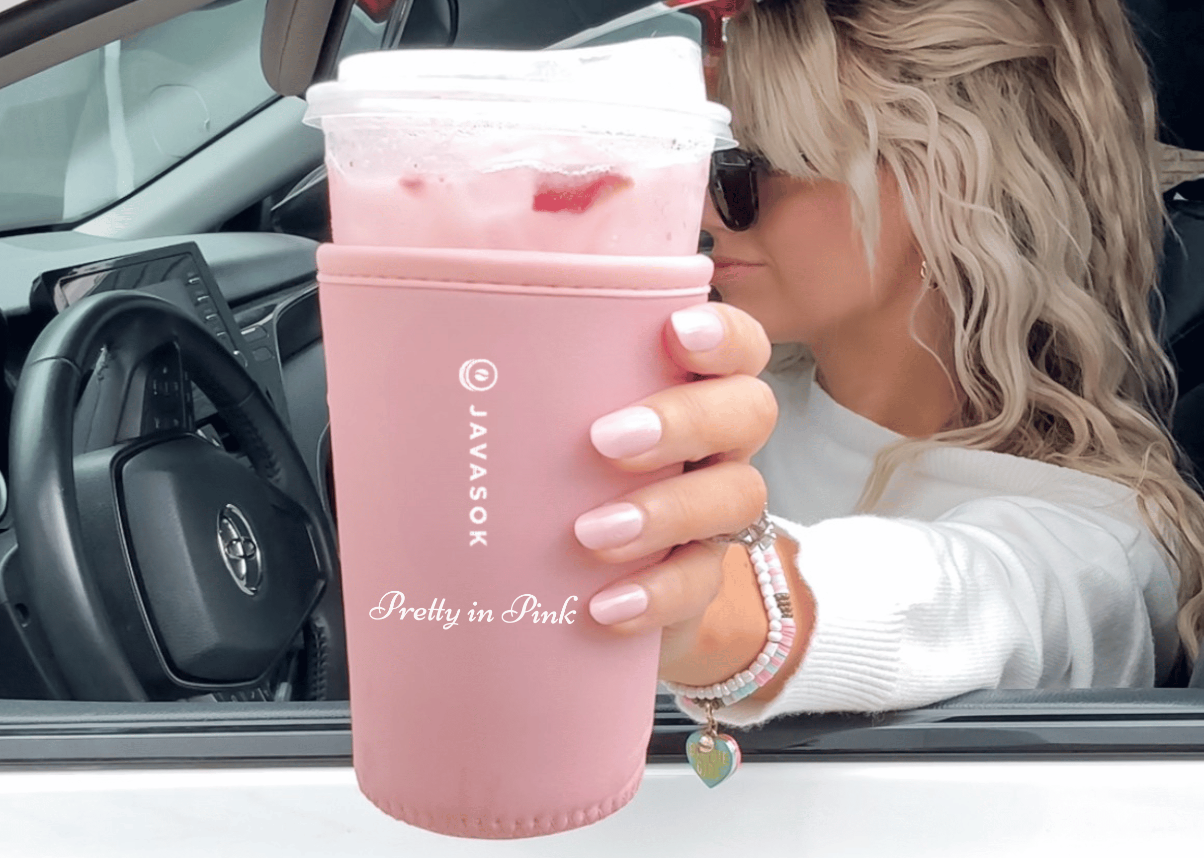 A woman in a white sweater and sunglasses holds a strawberry refresher with a pink insulated sleeve while sitting in a car. The sleeve is personalized with the phrase 'Pretty in Pink' 