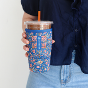 A person in a navy shirt and light blue jeans holds a blue floral insulated iced coffee sleeve