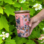 A hand holding a clear plastic tumbler with a floral neoprene sleeve in pink, navy, and peach tones, set against a background of green hydrangea leaves and white flowers.