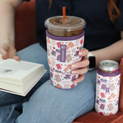 A person holding a clear plastic iced coffee cup with an orange straw, wrapped in a white reusable sleeve printed with purple and pink coffee and book-themed illustrations. A matching can cooler sits on their lap, and they are holding an open book.