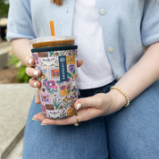 A person holding a clear plastic tumbler with a colorful floral and illustrated sleeve. The tumbler has a dark lid and an orange straw, and the person is wearing a light blue cardigan and blue jeans.