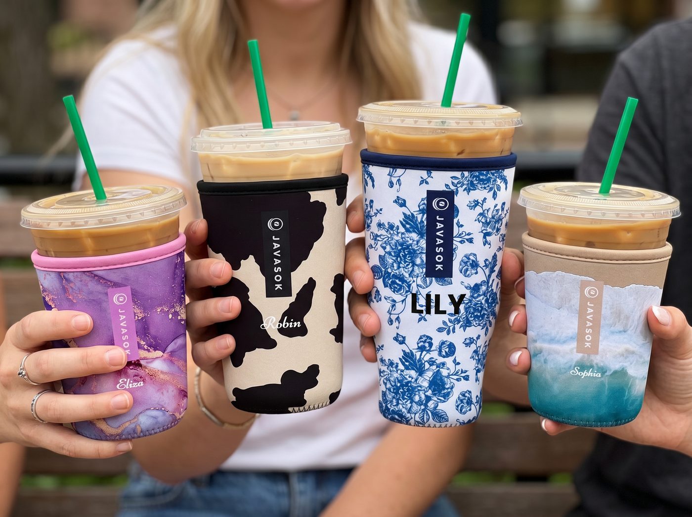 Four customized javasoks on iced coffee cups held up by people outdoors. The cups feature different colorful custom sleeves: purple marble, black and beige cow print, blue floral, and beige with a blue wave design.
